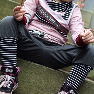 May include: A young person wearing a pink and black striped long-sleeved shirt with the word "photogenic" printed on it. They are also wearing black and white striped leggings and black high-top sneakers with pink accents. The person is sitting on a set of concrete steps.