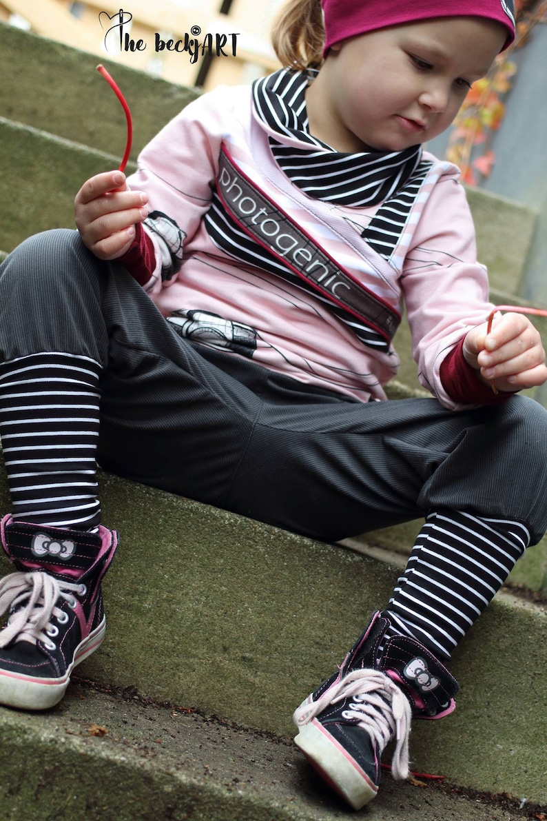 May include: A young person wearing a pink and black striped long-sleeved shirt with the word "photogenic" printed on it. They are also wearing black and white striped leggings and black high-top sneakers with pink accents. The person is sitting on a set of concrete steps.
