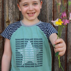 May include: A young boy wearing a green and blue striped shirt with the word "SCHULKIND" repeated on the front, smiles while holding a bouquet of pink and yellow flowers.