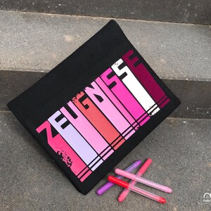 May include: Black zippered pouch with the word "ZEUGNISSE" printed in pink, red, and white stripes. The pouch is lying on a set of gray stairs with five pink and red markers in front of it.