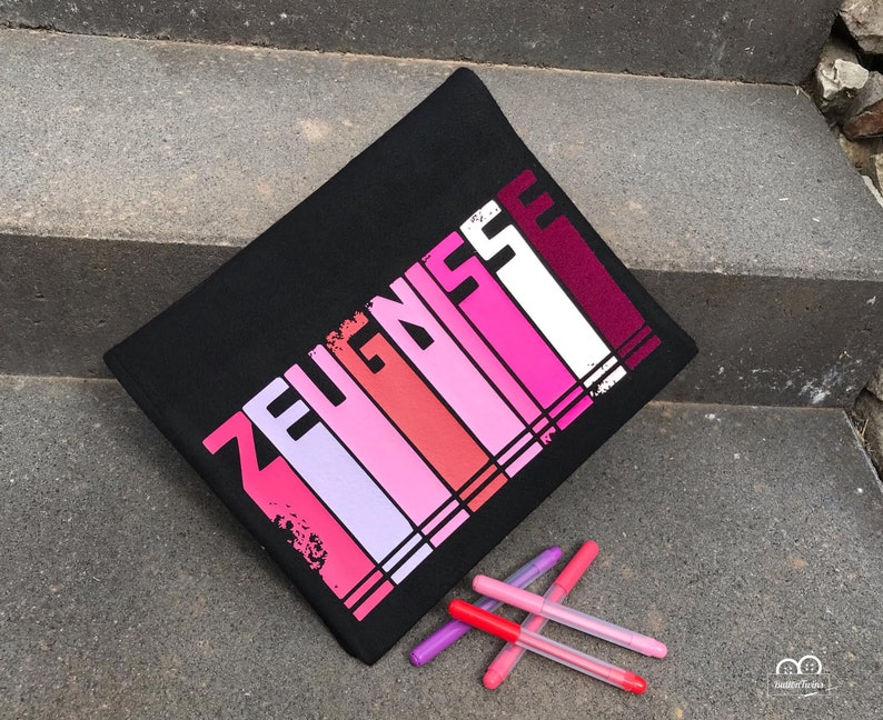 May include: Black zippered pouch with the word "ZEUGNISSE" printed in pink, red, and white stripes. The pouch is lying on a set of gray stairs with five pink and red markers in front of it.