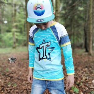 May include: A child wearing a blue and white long-sleeved shirt with a large black star and the letter "T" in the center. The shirt has a yellow trim around the neck and sleeves. The child is also wearing a blue baseball cap with a white shark graphic and the words "CHASED BY SHARKS SINCE 1994".