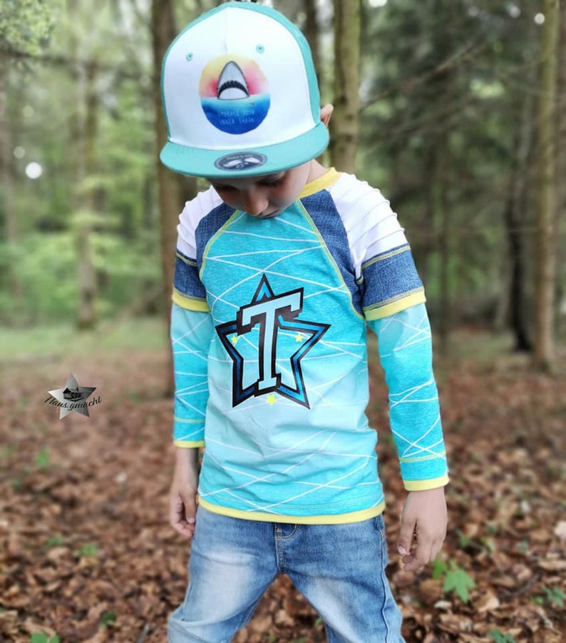 May include: A child wearing a blue and white long-sleeved shirt with a large black star and the letter "T" in the center. The shirt has a yellow trim around the neck and sleeves. The child is also wearing a blue baseball cap with a white shark graphic and the words "CHASED BY SHARKS SINCE 1994".