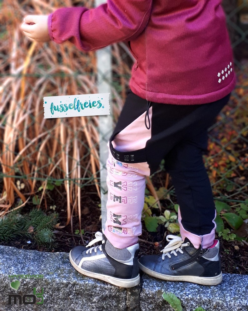 Puede incluir: Un ni&ntilde;o lleva una chaqueta y pantalones rosa y negro con la palabra "fusselfreies" en una etiqueta. Los pantalones tienen una raya rosa a un lado y el ni&ntilde;o lleva zapatillas negras y grises.