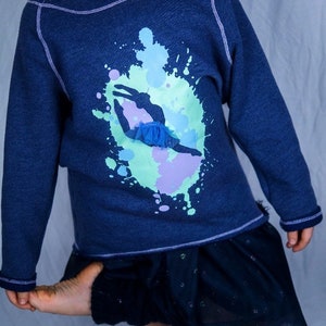 Op de afbeelding: Een donkerblauwe longsleeve met een afbeelding van een danseres in een blauwe tutu, omringd door kleurrijke verfspatten. De trui heeft een verweerde look en een paarse naad rond de halslijn.