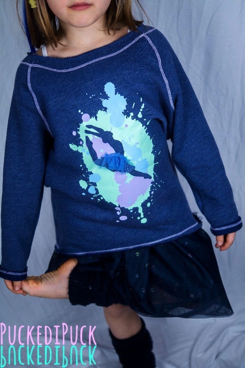 Op de afbeelding: Een donkerblauwe longsleeve met een afbeelding van een danseres in een blauwe tutu, omringd door kleurrijke verfspatten. De trui heeft een verweerde look en een paarse naad rond de halslijn.