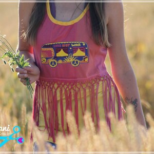 May include: A woman wearing a pink tank top with a fringe hem and a yellow graphic of a van with the text "Love Not War". She is standing in a field of tall grass and holding a bouquet of red poppies.