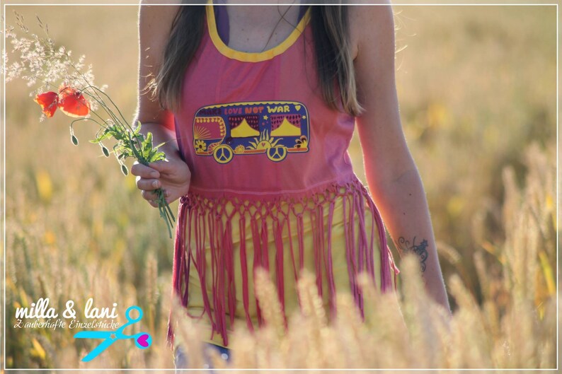 May include: A woman wearing a pink tank top with a fringe hem and a yellow graphic of a van with the text "Love Not War". She is standing in a field of tall grass and holding a bouquet of red poppies.