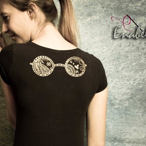 May include: A black t-shirt with a gold-colored design of a pair of round glasses with intricate details on the lenses. The glasses are centered on the back of the shirt.