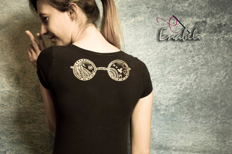May include: A black t-shirt with a gold-colored design of a pair of round glasses with intricate details on the lenses. The glasses are centered on the back of the shirt.