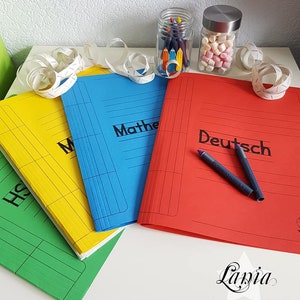 May include: Three colorful folders with the words "History", "Mathe", and "Deutsch" written on them. The folders are on a white surface with a jar of crayons and a jar of marshmallows in the background.