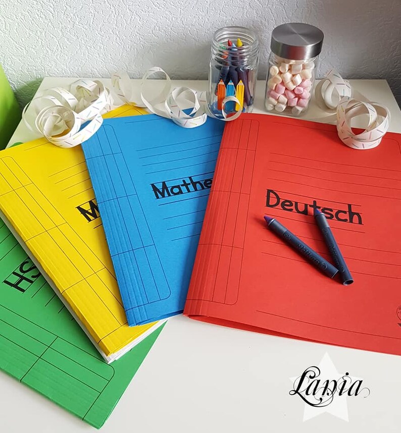May include: Three colorful folders with the words "History", "Mathe", and "Deutsch" written on them. The folders are on a white surface with a jar of crayons and a jar of marshmallows in the background.