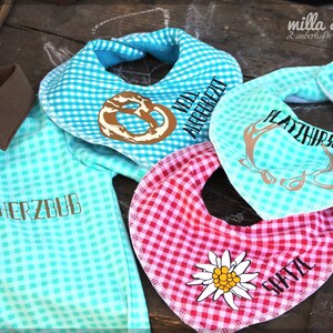 May include: A collection of baby items, including a turquoise and brown collared shirt and three bibs. The bibs are in turquoise and red gingham patterns, with various designs and text, including a pretzel, deer antlers, and a flower.