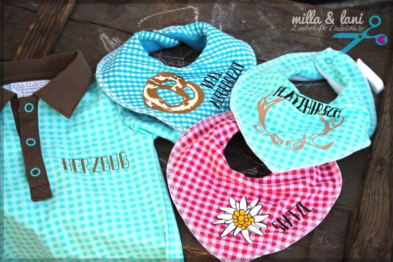 May include: A collection of baby items, including a turquoise and brown collared shirt and three bibs. The bibs are in turquoise and red gingham patterns, with various designs and text, including a pretzel, deer antlers, and a flower.