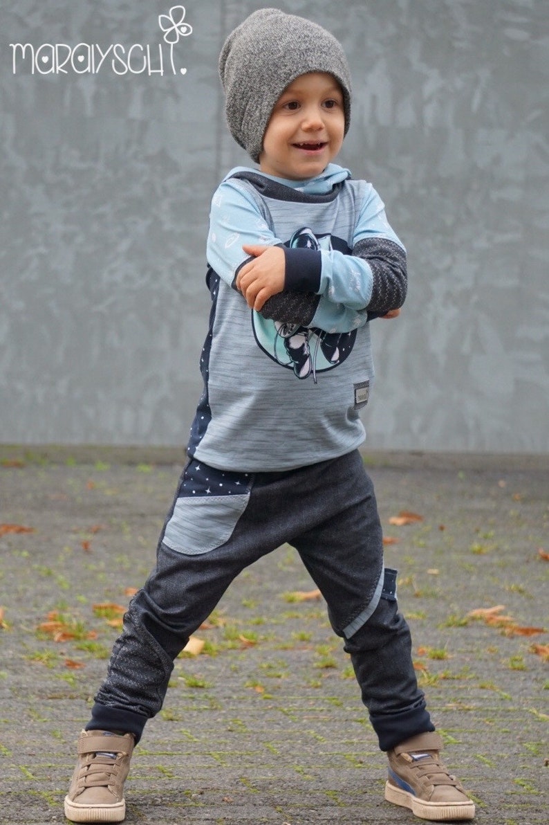 Puede incluir: Un ni&ntilde;o peque&ntilde;o con una sudadera con capucha azul y gris con un gr&aacute;fico de tem&aacute;tica espacial, gorro gris, jeans azul oscuro con un bolsillo de parche y zapatillas marrones.