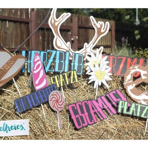 May include: A collection of Oktoberfest-themed photo booth props on sticks, including a cowboy hat, antlers, a pretzel, and various word cutouts. The props are arranged on a bed of hay, with a blurred background.