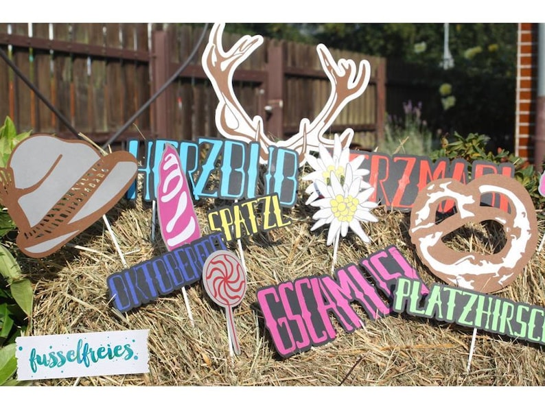 May include: A collection of Oktoberfest-themed photo booth props on sticks, including a cowboy hat, antlers, a pretzel, and various word cutouts. The props are arranged on a bed of hay, with a blurred background.