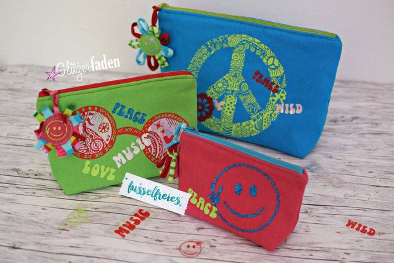 May include: Three colorful zippered pouches with peace signs, smiley faces, and the words "Peace Music Love" and "Peace Wild" printed on them. The pouches are made of fabric and have a zipper closure.