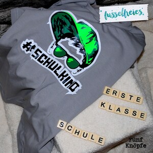 May include: Gray t-shirt with a green and black graphic of a person wearing sunglasses and a hat. The text "#SCHULKIND" is on the shirt. The words "ERSTE KLASSE" and "SCHULE" are spelled out with letter tiles.