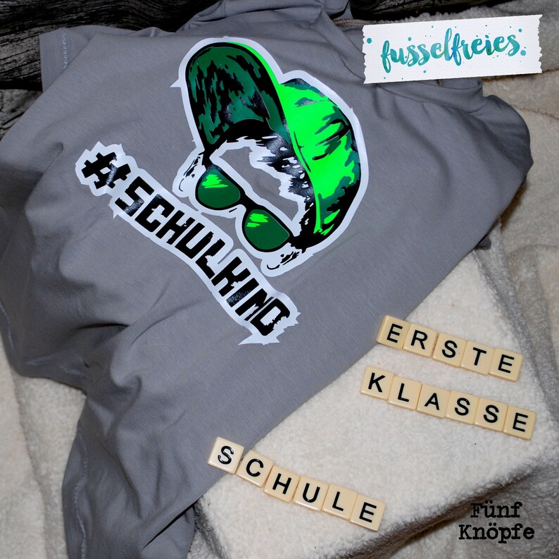May include: Gray t-shirt with a green and black graphic of a person wearing sunglasses and a hat. The text "#SCHULKIND" is on the shirt. The words "ERSTE KLASSE" and "SCHULE" are spelled out with letter tiles.