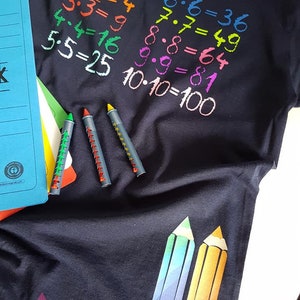 May include: A black t-shirt with colorful numbers and multiplication equations printed on the front. The equations are 1x1=1, 2x2=4, 3x3=9, 4x4=16, 5x5=25, 6x6=36, 7x7=49, 8x8=64, 9x9=81, and 10x10=100. There are three colorful crayons on the left side of the shirt.