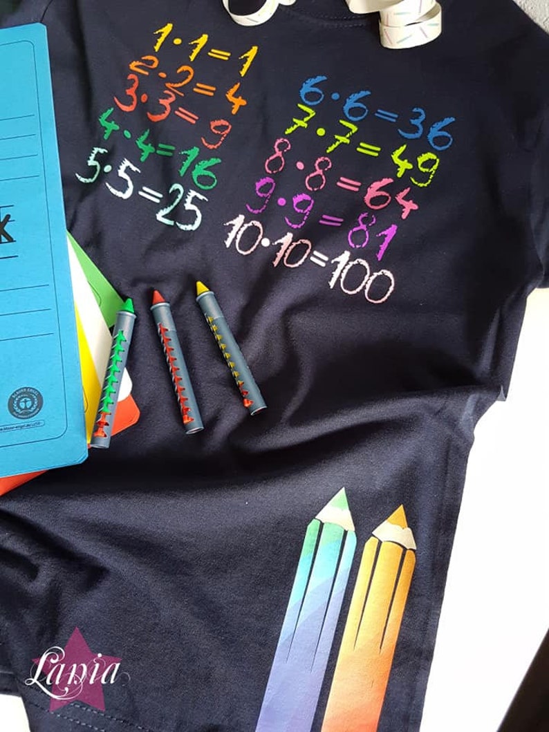May include: A black t-shirt with colorful numbers and multiplication equations printed on the front. The equations are 1x1=1, 2x2=4, 3x3=9, 4x4=16, 5x5=25, 6x6=36, 7x7=49, 8x8=64, 9x9=81, and 10x10=100. There are three colorful crayons on the left side of the shirt.
