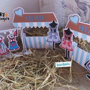 May include: A whimsical paper craft scene featuring miniature clothing displayed on a clothesline and mannequins. The scene includes a market stall with blue and white striped accents, and the text "Art of Singer". The clothing includes dresses and shorts.