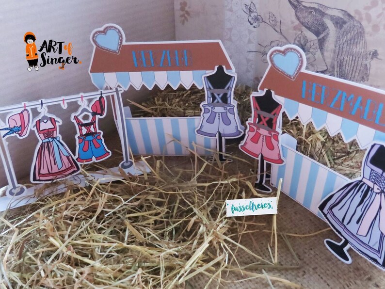 May include: A whimsical paper craft scene featuring miniature clothing displayed on a clothesline and mannequins. The scene includes a market stall with blue and white striped accents, and the text "Art of Singer". The clothing includes dresses and shorts.