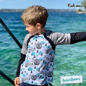 May include: A child wearing a long-sleeved shirt with a black and white pattern of Viking helmets and blue and white geometric shapes. The shirt has black sleeves and a gray and black raglan sleeve. The child is standing in front of a body of water.