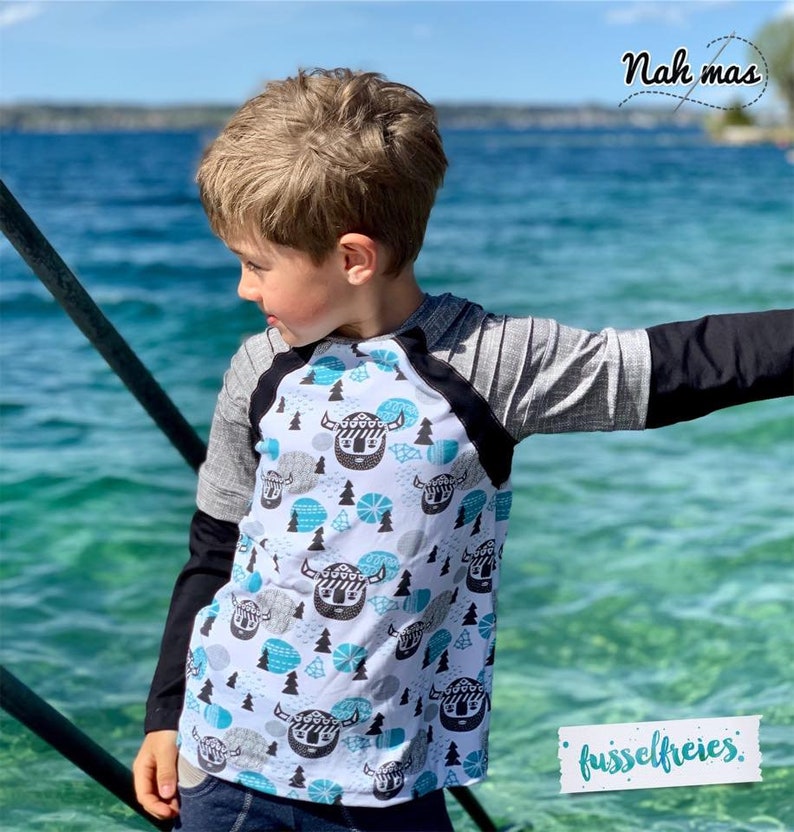 May include: A child wearing a long-sleeved shirt with a black and white pattern of Viking helmets and blue and white geometric shapes. The shirt has black sleeves and a gray and black raglan sleeve. The child is standing in front of a body of water.