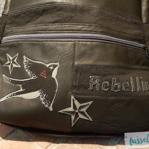May include: Black leather bag with a zippered pocket and embroidered details. The bag features a white swallow with a red heart, two white stars, and the word "Rebellion" in white thread.