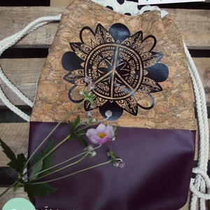 May include: A drawstring bag made of cork with a black and white peace sign mandala design. The bag has a purple bottom and a white drawstring. The text "fusselfreies" and "ZENTI MADCHEN" are visible on the bag.