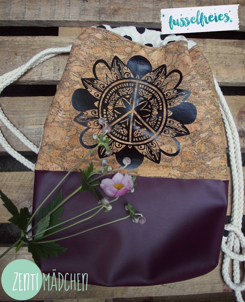 May include: A drawstring bag made of cork with a black and white peace sign mandala design. The bag has a purple bottom and a white drawstring. The text "fusselfreies" and "ZENTI MADCHEN" are visible on the bag.