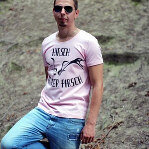 May include: A person wearing a light pink t-shirt with the text "HIRSCH AUF DER PIRSCH" and a graphic of antlers. The person is also wearing blue jeans and sunglasses. The image also includes the text "Isa's N&auml;hwelt & Kreativhund".