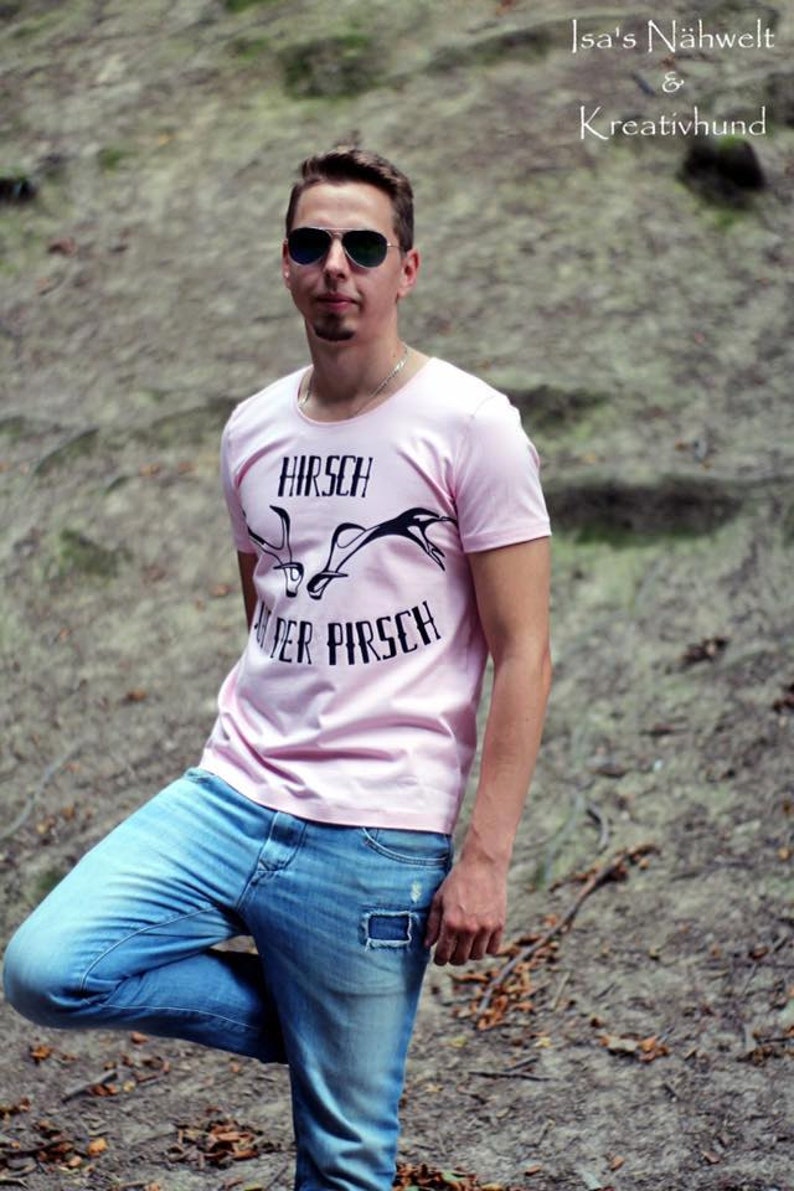 May include: A person wearing a light pink t-shirt with the text "HIRSCH AUF DER PIRSCH" and a graphic of antlers. The person is also wearing blue jeans and sunglasses. The image also includes the text "Isa's N&auml;hwelt & Kreativhund".