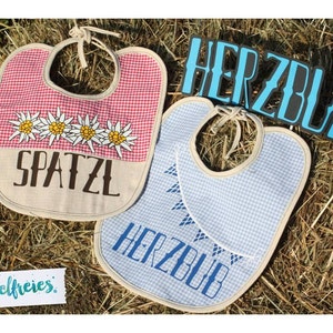 May include: Two baby bibs with a Bavarian theme. One bib is red and white checkered with the word "SPATZL" and edelweiss flowers. The other is blue and white checkered with the word "HERZBUB". Both have tie closures.