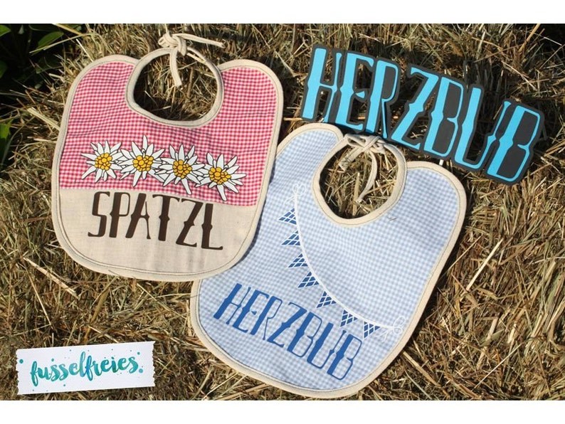 May include: Two baby bibs with a Bavarian theme. One bib is red and white checkered with the word "SPATZL" and edelweiss flowers. The other is blue and white checkered with the word "HERZBUB". Both have tie closures.