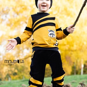 May include: A young child wearing a yellow and black striped sweater with a raccoon graphic and black pants with yellow stripes. The child is holding a stick and smiling.