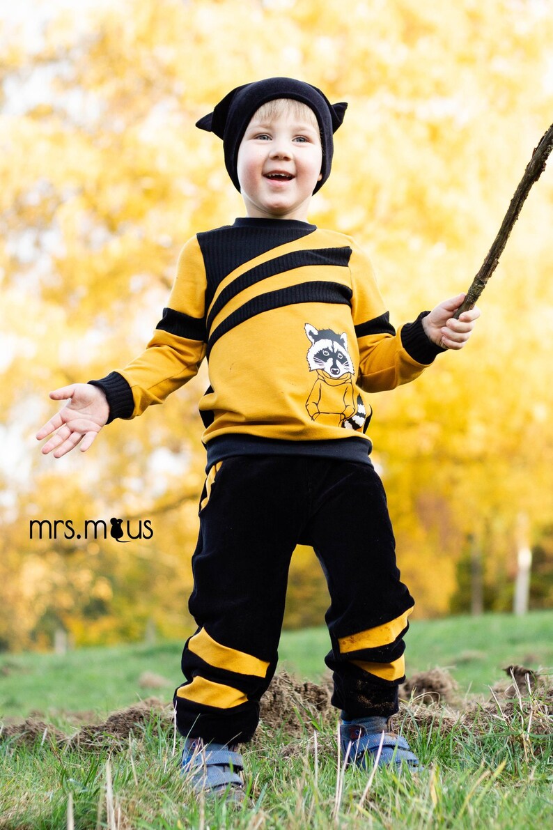 May include: A young child wearing a yellow and black striped sweater with a raccoon graphic and black pants with yellow stripes. The child is holding a stick and smiling.