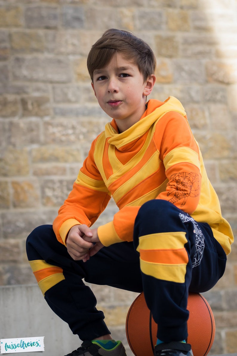 May include: A young person wearing a bright orange and yellow striped hoodie with a black and yellow striped waistband. The person is sitting on a basketball.