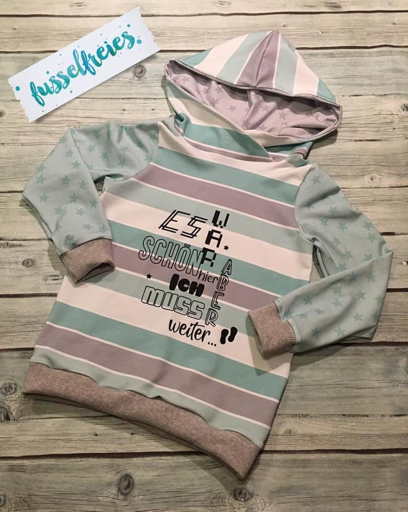 May include: A light blue and gray striped hooded sweatshirt with a star pattern on the sleeves. The sweatshirt has a text design that reads "Es war sch&ouml;n, aber ich muss weiter..."