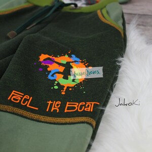 Op de afbeelding: Groene en lichtgroene kinderjoggingbroek met een kleurrijke afbeelding van een springend persoon en de tekst "Feel the Beat" in oranje. De joggingbroek heeft een geel trekkoord en de merknaam "Jakoki" staat aan de zijkant gedrukt.