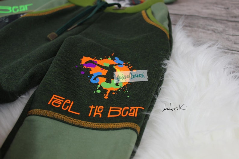 Op de afbeelding: Groene en lichtgroene kinderjoggingbroek met een kleurrijke afbeelding van een springend persoon en de tekst "Feel the Beat" in oranje. De joggingbroek heeft een geel trekkoord en de merknaam "Jakoki" staat aan de zijkant gedrukt.