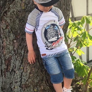 May include: A young boy wearing a grey and black baseball cap with the letters "NY" on it. He is wearing a white t-shirt with a blue and white striped design and a ship graphic. He is also wearing blue denim shorts and white sneakers.