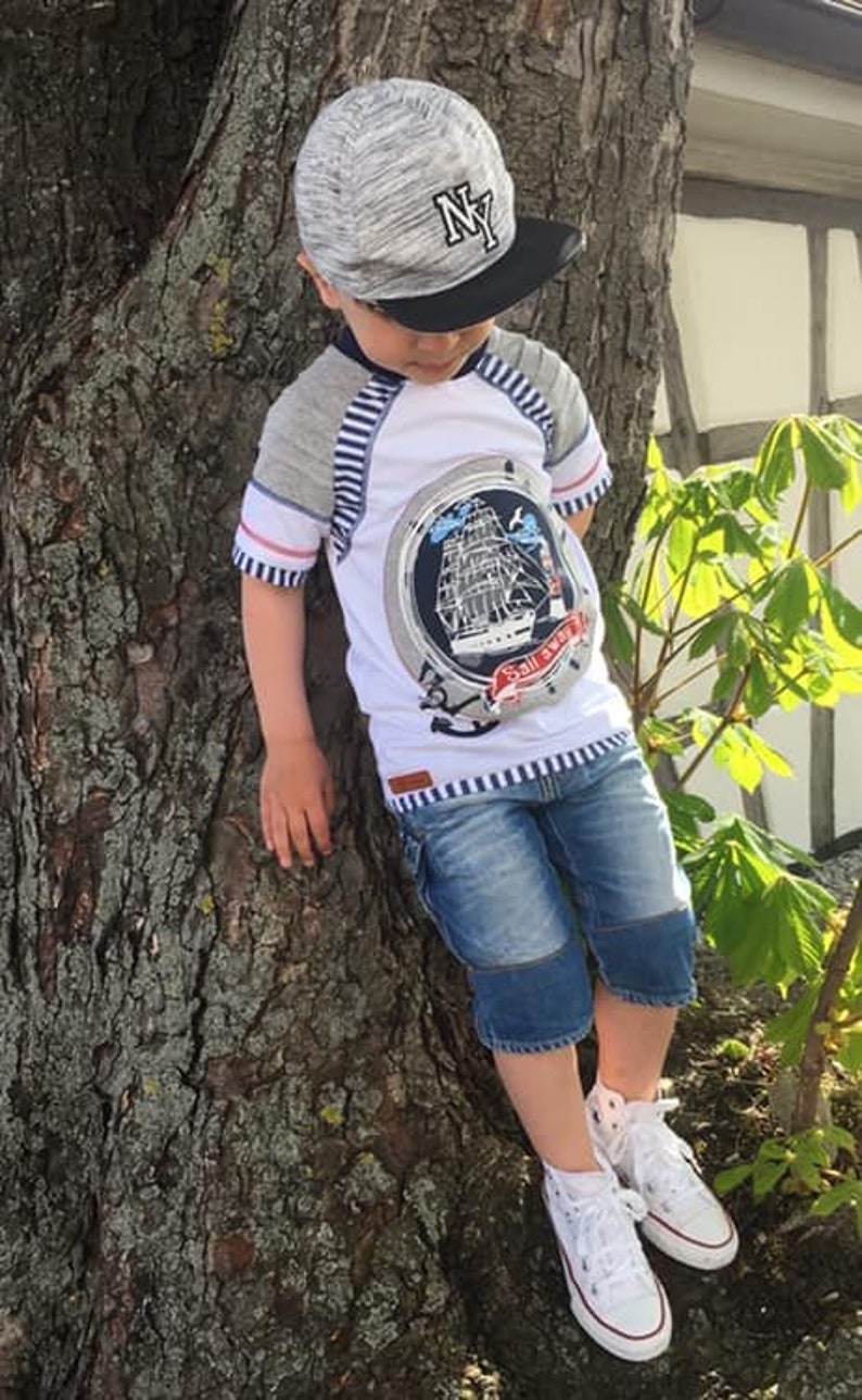 May include: A young boy wearing a grey and black baseball cap with the letters "NY" on it. He is wearing a white t-shirt with a blue and white striped design and a ship graphic. He is also wearing blue denim shorts and white sneakers.