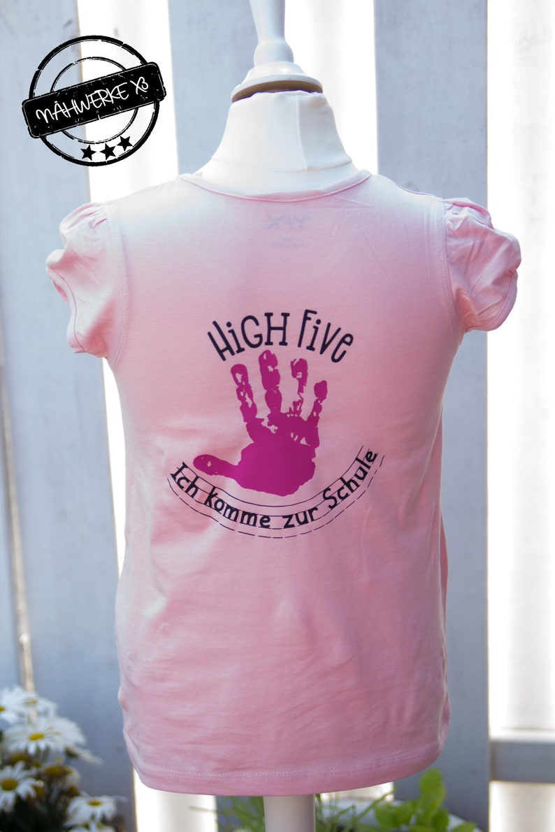 May include: Pink t-shirt with a black and pink design that says "HIGH Five Ich komme zur Schule".