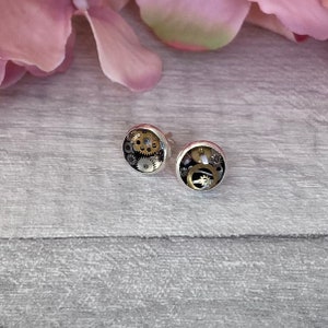 May include: A pair of silver stud earrings with a round, clear resin design featuring a cluster of gold and silver gears.