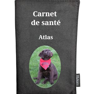 May include: A black fabric health record cover featuring the white text Carnet de santé and Atlas above an oval photograph of a brown puppy wearing a red bandana. A small black tag with the white text KIPAPEE is attached to the side.