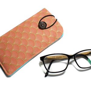 May include: A coral-colored glasses case with a gold and peach fan pattern and a blue trim. The case has a black elastic strap with a star-shaped button. A pair of black framed glasses with a wooden bamboo arm is lying on a white surface.