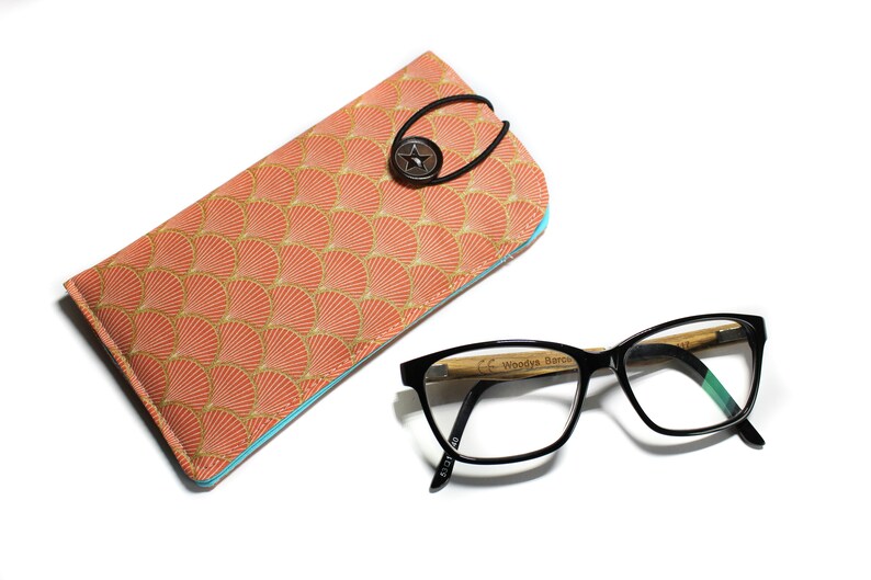 May include: A coral-colored glasses case with a gold and peach fan pattern and a blue trim. The case has a black elastic strap with a star-shaped button. A pair of black framed glasses with a wooden bamboo arm is lying on a white surface.